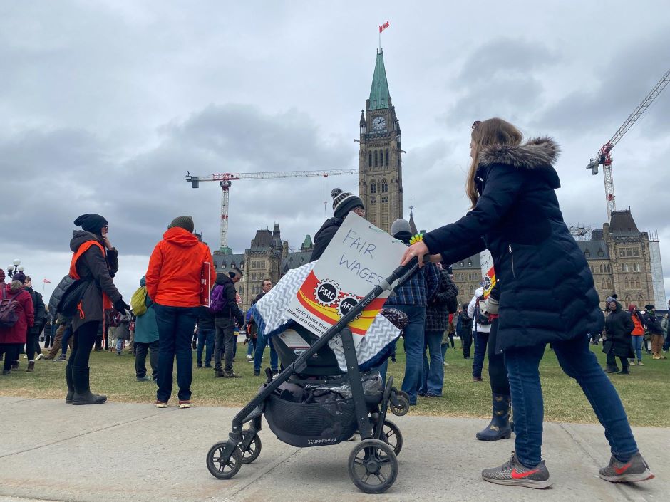 A worker pushes a stroller in front of Parliament Hill with a sign saying "fair wages."